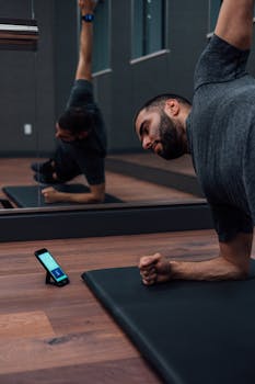 A bearded man stretching on a yoga mat while using a smartphone app in a gym studio with mirrors.