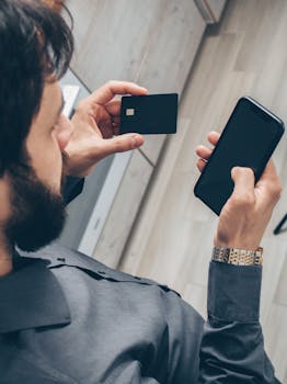 Adult man using a smartphone for online transactions while holding a credit card indoors.