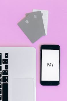 Top view of mobile payment setup with smartphone, credit cards, and laptop on pink background.