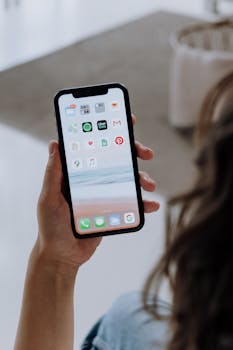 A person holding a smartphone displaying various application icons in an indoor setting.