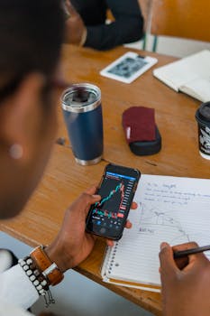 A person reviewing stock market data on a smartphone while taking notes during a meeting.