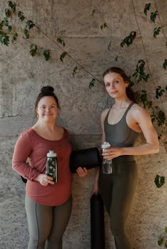 Two women holding yoga mats and water bottles, ready for indoor yoga practice.