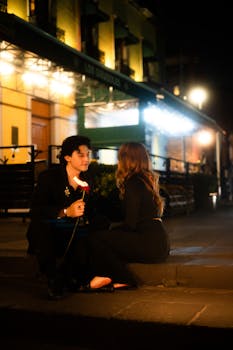 A couple shares a romantic moment sitting on a nighttime street in Mexico City, holding a rose.
