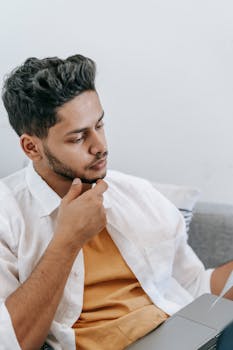 A pensive man sitting on a couch, focused on his laptop in a cozy home setting.